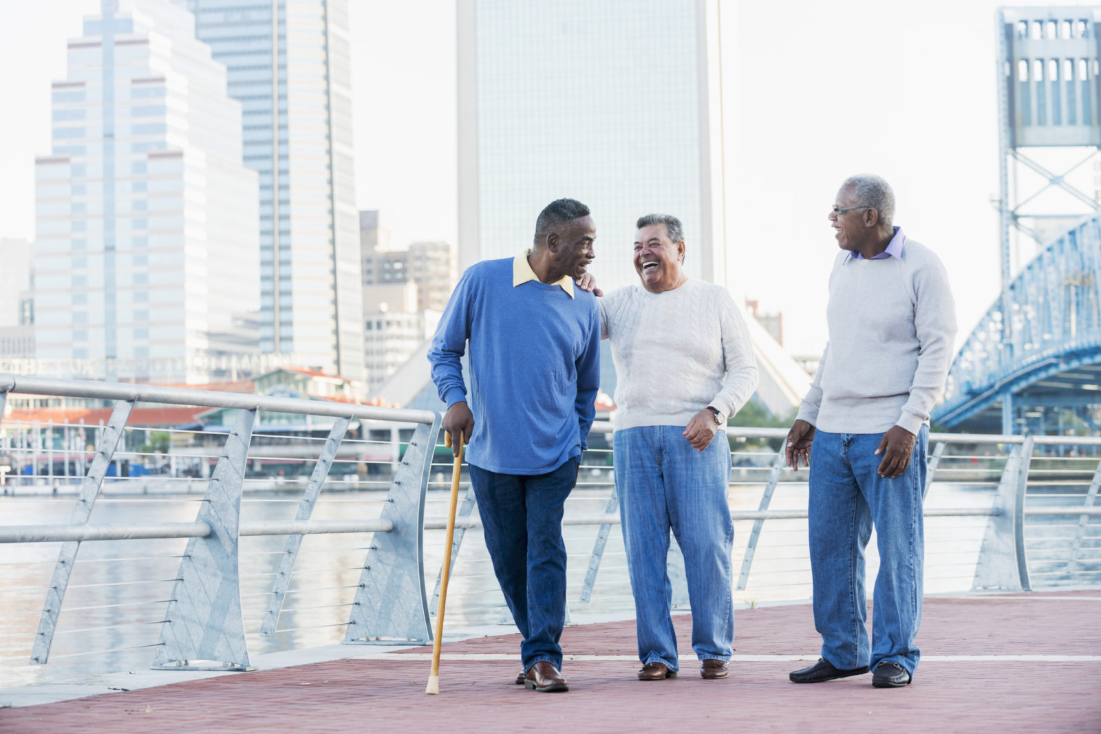 A group of three multi-ethnic senior men walking together along a city waterfront, laughing and talking.